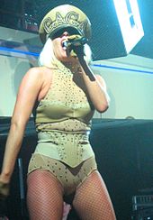 A blond woman standing. She is wearing a khaki colored leotard and an admirals cap. She is holding a microphone to her mouth in her left hand.