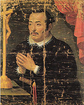 Pintura de hombre asiático con alzacuellos y sotana negra rezando frente a un crucifijo.