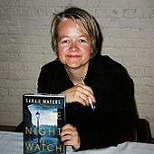 Image of a women with short blond hair seated at a table holding a pen and a book cover to the camera