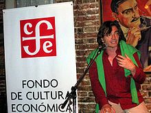  Head and shoulders of a man in his early fifties, with the logo of a publishing house and the painting of an Argentinian artist in his back. The man is speaking into a microphone, and gesturing.