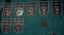 Seven large black shield-shaped banners are hung on a green wall, with white text for the name and number, or role that the individual played.