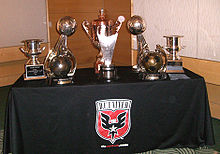 A table holding seven golden trophies of various sizes. The table is cover by a cloth with the team's shield on it.