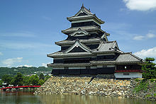 Torre de castillo de cinco plantas con muros de madera negra, ubicada sobre una plataforma de piedras sin tallar y ods lados rodeados por agua. La torre está conectada a la estructura inferior.