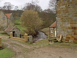 A small stream flowing under a bridge, flanked on either side by grey stone buildings.