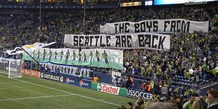 Fans in a stadium display two large banners which read 'The Boys From Seattle Are Back' with artwork of soccer players in green and blue uniforms.